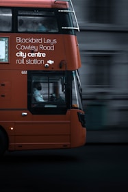 An orange double-decker bus in motion with destination details like Blackbird Leys, Cowley Road, city centre, and rail station displayed on its side. The focal point is the bus driver visible through the large window. The background appears blurred, emphasizing the bus's movement.