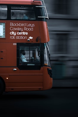 An orange double-decker bus in motion with destination details like Blackbird Leys, Cowley Road, city centre, and rail station displayed on its side. The focal point is the bus driver visible through the large window. The background appears blurred, emphasizing the bus's movement.