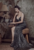 A woman in an elegant black sequined gown sits on a woven stool. Her hair is styled in vintage waves, and she is posed gracefully with one hand touching her chin. The setting includes an antique telephone, a decorative vase, and pampas grass, all on a small wooden table. A vintage lamp adds a classic touch to the scene.