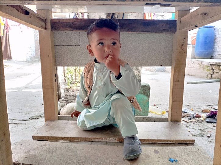 A young child wearing traditional clothing sits under a wooden structure. The child has a thoughtful expression, with one finger touching their lips. The setting appears to be outdoors with scattered objects on the ground and a large blue container in the background.