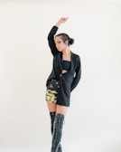 A person poses with one arm raised, wearing a black blazer and a short skirt with a dragon print. They have knee-high black lace-up boots and bold makeup with dark eyeliner. The background is light and plain, highlighting the stylish and edgy outfit.