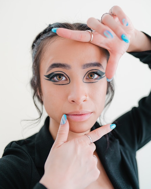 A close-up of a person using their hands to frame their face, emphasizing dramatic eye makeup and unique nail art. The person has bold eyeliner and eyeshadow, with light blue and white striped nails. They are wearing a black top and have multiple rings on their fingers.