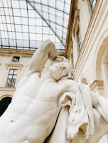 A classical marble statue depicting a muscular figure with a serene expression, resting one arm behind its head. The sculpture is set against a backdrop of a grand building with large windows and a glass ceiling, indicating an indoor location such as a museum.