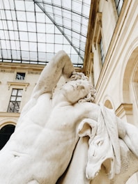 A classical marble statue depicting a muscular figure with a serene expression, resting one arm behind its head. The sculpture is set against a backdrop of a grand building with large windows and a glass ceiling, indicating an indoor location such as a museum.