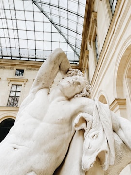 A classical marble statue depicting a muscular figure with a serene expression, resting one arm behind its head. The sculpture is set against a backdrop of a grand building with large windows and a glass ceiling, indicating an indoor location such as a museum.