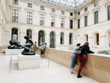 Two sculptures are prominently displayed in a spacious gallery with a high glass ceiling. Several people are engaging with the art and surroundings, leaning against the railing and observing. The architecture features tall arched windows set in a classical style.