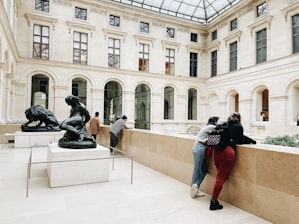 Two sculptures are prominently displayed in a spacious gallery with a high glass ceiling. Several people are engaging with the art and surroundings, leaning against the railing and observing. The architecture features tall arched windows set in a classical style.