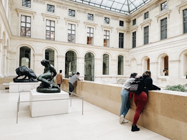Two sculptures are prominently displayed in a spacious gallery with a high glass ceiling. Several people are engaging with the art and surroundings, leaning against the railing and observing. The architecture features tall arched windows set in a classical style.