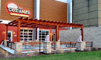 A restaurant exterior with a large wooden pergola adorned with lights, flanked by two lion statues on stone pedestals. The building features a colorful sign labeled 'Mi Cozumel' and offers margaritas and flaming fajitas. Outdoor seating is arranged under the pergola, surrounded by a metal railing and a small landscaped area with grass and dried plants.