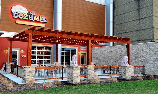 A restaurant exterior with a large wooden pergola adorned with lights, flanked by two lion statues on stone pedestals. The building features a colorful sign labeled 'Mi Cozumel' and offers margaritas and flaming fajitas. Outdoor seating is arranged under the pergola, surrounded by a metal railing and a small landscaped area with grass and dried plants.