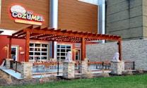 A restaurant exterior with a large wooden pergola adorned with lights, flanked by two lion statues on stone pedestals. The building features a colorful sign labeled 'Mi Cozumel' and offers margaritas and flaming fajitas. Outdoor seating is arranged under the pergola, surrounded by a metal railing and a small landscaped area with grass and dried plants.