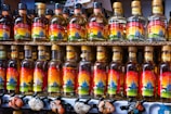 Multiple bottles of mezcal neatly arranged on shelves, each with a colorful label featuring an agave plant and various designs against a sunset backdrop. Small animal figurines are displayed beneath the bottles.