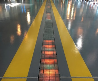 A shiny floor surface features two parallel yellow lines, with reflections of colorful lights scattered across it. In the center between the yellow lines, a gridded pattern runs along the ground, illuminated with orange and red hues.
