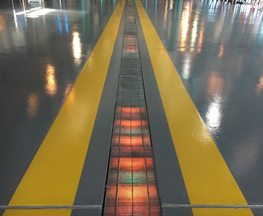 A shiny floor surface features two parallel yellow lines, with reflections of colorful lights scattered across it. In the center between the yellow lines, a gridded pattern runs along the ground, illuminated with orange and red hues.
