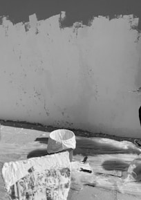 A section of a wall is being painted. The surface appears partially covered with paint, and various painting supplies are present, including a large paint bucket and a brush. The ground is protected with plastic sheets, and there is a close-up of paint on a tool in the foreground.