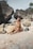 A woman wearing a floral bikini sits on a sandy beach surrounded by large rocks. She appears relaxed with eyes closed in a sunbathing position. The natural setting and scattered boulders create a serene atmosphere.