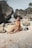 A woman wearing a floral bikini sits on a sandy beach surrounded by large rocks. She appears relaxed with eyes closed in a sunbathing position. The natural setting and scattered boulders create a serene atmosphere.
