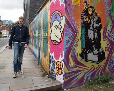 A man wearing a dark jacket and jeans is walking along an urban street. The wall beside him is brightly decorated with colorful graffiti art, including designs of faces and abstract patterns. The graffiti features vibrant colors and intricate designs, contributing to a lively urban atmosphere.