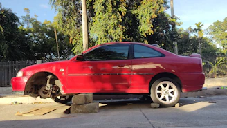 A red car with one of its front wheels missing is propped up on concrete blocks on a street, surrounded by lush green trees and a metal fence.