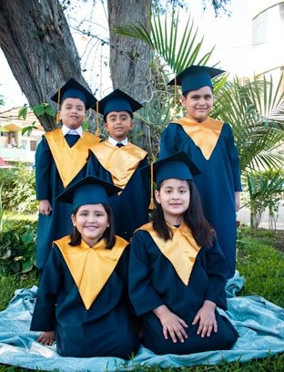 a group of children in graduation gowns sitting under a tree