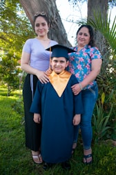 A young child wearing a graduation cap and gown stands smiling, flanked by two women, presumed to be family members. They are standing outdoors in a grassy area with a large tree and some plants in the background. One woman is wearing a purple top and black skirt while the other has a floral patterned shirt and jeans. The group appears happy and proud as they celebrate an educational milestone.