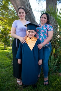 A young child wearing a graduation cap and gown stands smiling, flanked by two women, presumed to be family members. They are standing outdoors in a grassy area with a large tree and some plants in the background. One woman is wearing a purple top and black skirt while the other has a floral patterned shirt and jeans. The group appears happy and proud as they celebrate an educational milestone.