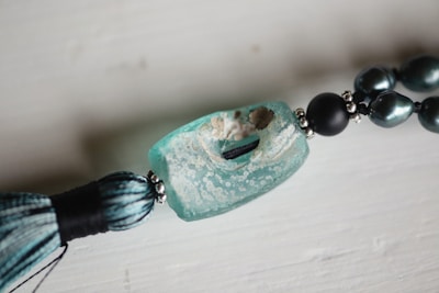 A close-up view of a decorative piece featuring a large, rectangular turquoise bead with a textured surface, paired with a tassel and dark, metallic beads strung together. The main bead has a rugged, weathered appearance with visible holes and a speckled pattern.