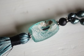 A close-up view of a decorative piece featuring a large, rectangular turquoise bead with a textured surface, paired with a tassel and dark, metallic beads strung together. The main bead has a rugged, weathered appearance with visible holes and a speckled pattern.
