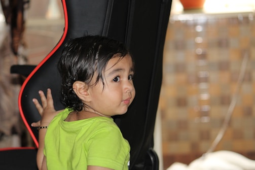 A young child with dark, wet hair is leaning against a black chair with a red outline, looking behind them. The child is wearing a bright green shirt, and a bracelet on their wrist is visible. The background features a blurred mosaic pattern with neutral tones.