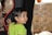 A young child with dark, wet hair is leaning against a black chair with a red outline, looking behind them. The child is wearing a bright green shirt, and a bracelet on their wrist is visible. The background features a blurred mosaic pattern with neutral tones.