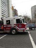 A red and white fire truck is moving quickly through an urban street intersection. The fire truck has a P6 designation on its side and is equipped with emergency cones and firefighting equipment. Surrounding the fire truck are tall buildings and trees, creating an urban backdrop. Nearby, a blue SUV is visible on the road.