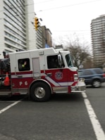 A red and white fire truck is moving quickly through an urban street intersection. The fire truck has a P6 designation on its side and is equipped with emergency cones and firefighting equipment. Surrounding the fire truck are tall buildings and trees, creating an urban backdrop. Nearby, a blue SUV is visible on the road.