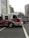 A red and white fire truck is moving quickly through an urban street intersection. The fire truck has a P6 designation on its side and is equipped with emergency cones and firefighting equipment. Surrounding the fire truck are tall buildings and trees, creating an urban backdrop. Nearby, a blue SUV is visible on the road.