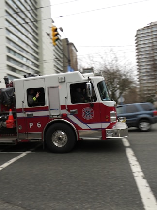 A red and white fire truck is moving quickly through an urban street intersection. The fire truck has a P6 designation on its side and is equipped with emergency cones and firefighting equipment. Surrounding the fire truck are tall buildings and trees, creating an urban backdrop. Nearby, a blue SUV is visible on the road.