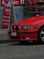 A red BMW car is parked near a roadside with a 'Dollar' shop sign in the background. The shop sign shows colorful decorative pots stacked on shelves. The pavement is visible below the car.