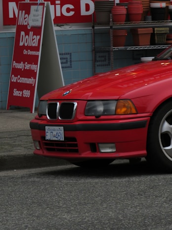A red BMW car is parked near a roadside with a 'Dollar' shop sign in the background. The shop sign shows colorful decorative pots stacked on shelves. The pavement is visible below the car.