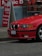 A red BMW car is parked near a roadside with a 'Dollar' shop sign in the background. The shop sign shows colorful decorative pots stacked on shelves. The pavement is visible below the car.