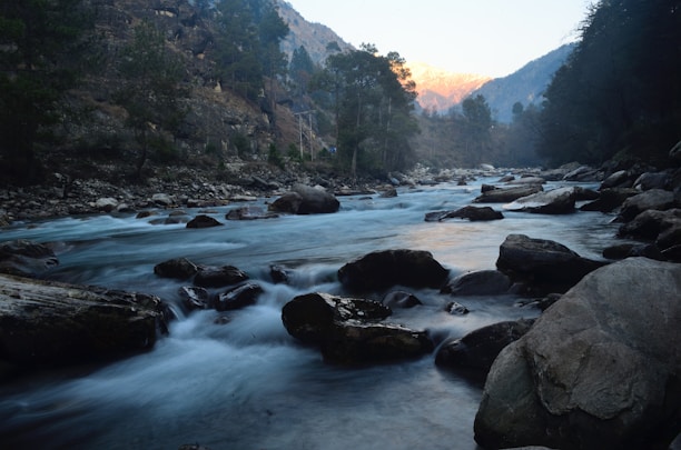 A serene river flowing through rocky terrain with mountains in the background at sunset.