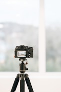 A professional camera is mounted on a tripod indoors, facing a large window with a blurred view of the outside. The camera's display screen shows a focused image, likely a landscape or architectural scene.