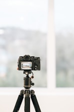 A professional camera is mounted on a tripod indoors, facing a large window with a blurred view of the outside. The camera's display screen shows a focused image, likely a landscape or architectural scene.