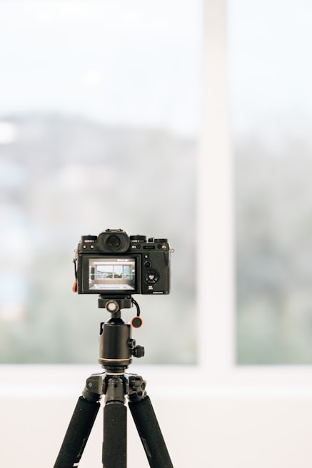 A professional camera is mounted on a tripod indoors, facing a large window with a blurred view of the outside. The camera's display screen shows a focused image, likely a landscape or architectural scene.