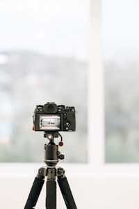 A professional camera is mounted on a tripod indoors, facing a large window with a blurred view of the outside. The camera's display screen shows a focused image, likely a landscape or architectural scene.