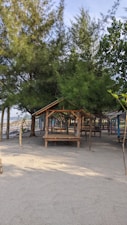 A peaceful beach setting with several wooden huts resting among tall, green trees. The sandy ground is smooth and the area looks calm and inviting. The sky is partly cloudy, giving a relaxed vibe.
