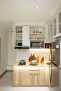 A modern, compact kitchen setup with wooden cabinets and white countertops. Open shelves display neatly stacked dishes and glassware. A small stove with a pot is on the counter, next to a cutting board and a set of knives. Above the counter, a microwave is built into the cabinet space. The wall features a white hexagonal tile pattern. The refrigerator is stainless steel, and various containers are placed on the shelves.