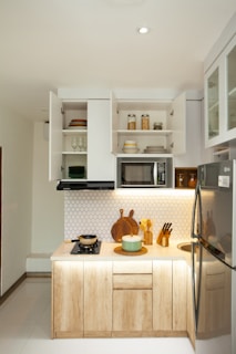 A modern, compact kitchen setup with wooden cabinets and white countertops. Open shelves display neatly stacked dishes and glassware. A small stove with a pot is on the counter, next to a cutting board and a set of knives. Above the counter, a microwave is built into the cabinet space. The wall features a white hexagonal tile pattern. The refrigerator is stainless steel, and various containers are placed on the shelves.