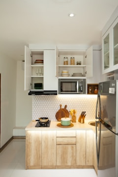 A modern, compact kitchen setup with wooden cabinets and white countertops. Open shelves display neatly stacked dishes and glassware. A small stove with a pot is on the counter, next to a cutting board and a set of knives. Above the counter, a microwave is built into the cabinet space. The wall features a white hexagonal tile pattern. The refrigerator is stainless steel, and various containers are placed on the shelves.