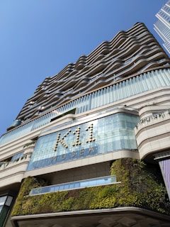 A modern building with a unique wavy architectural design and greenery integrated into its facade. The sign 'K11 MUSEA' is prominently displayed, and the structure contrasts against a clear blue sky.