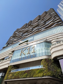A modern building with a unique wavy architectural design and greenery integrated into its facade. The sign 'K11 MUSEA' is prominently displayed, and the structure contrasts against a clear blue sky.