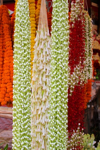 A vibrant array of floral garlands, featuring long strands of orange, yellow, white, and red flowers. The flowers are densely packed, creating thick, colorful lines with intricate textures.
