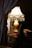 An elegantly decorated table lamp with a brass base and a cream-colored shade stands on a wooden table. The lamp emits a warm glow, illuminating the surrounding area. Crystal pendants hang from the edge of the lamp shade, adding a touch of sophistication. In the foreground, a thick curtain with a decorative tassel partially frames the scene. On the table, books are stacked neatly, suggesting a cozy and inviting space.